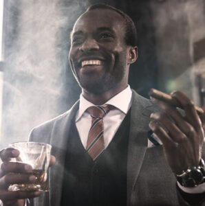 Man in a grey suit smiling, drinking and smoking a cigar